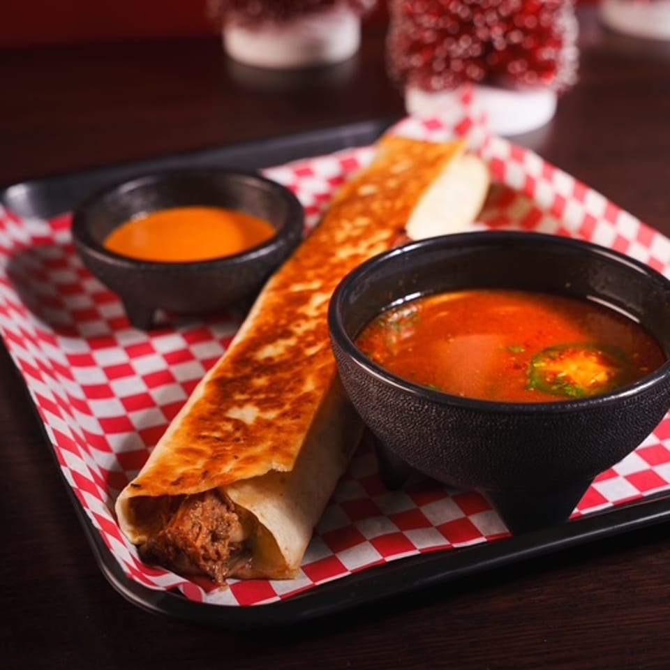 Crispy flauta, red soup, and orange dipping sauce on a checkered tray.