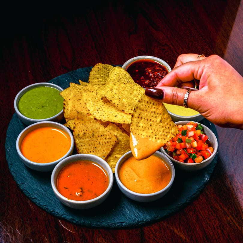 A hand dips a tortilla chip into a bowl of cheese sauce, surrounded by other dips.