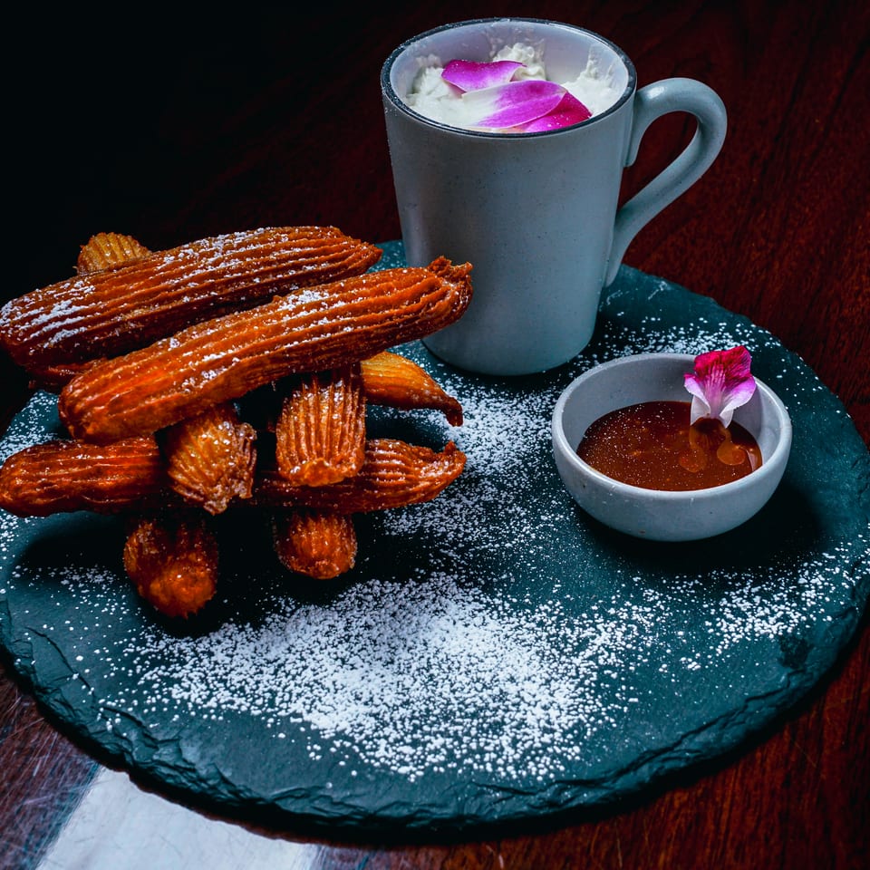 Golden churros dusted with powdered sugar, served with a dipping sauce and a creamy drink.