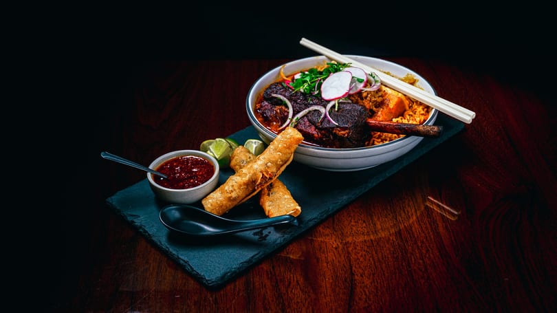 Braised short rib ramen with egg rolls, chili sauce, and lime wedges on a dark wooden table.